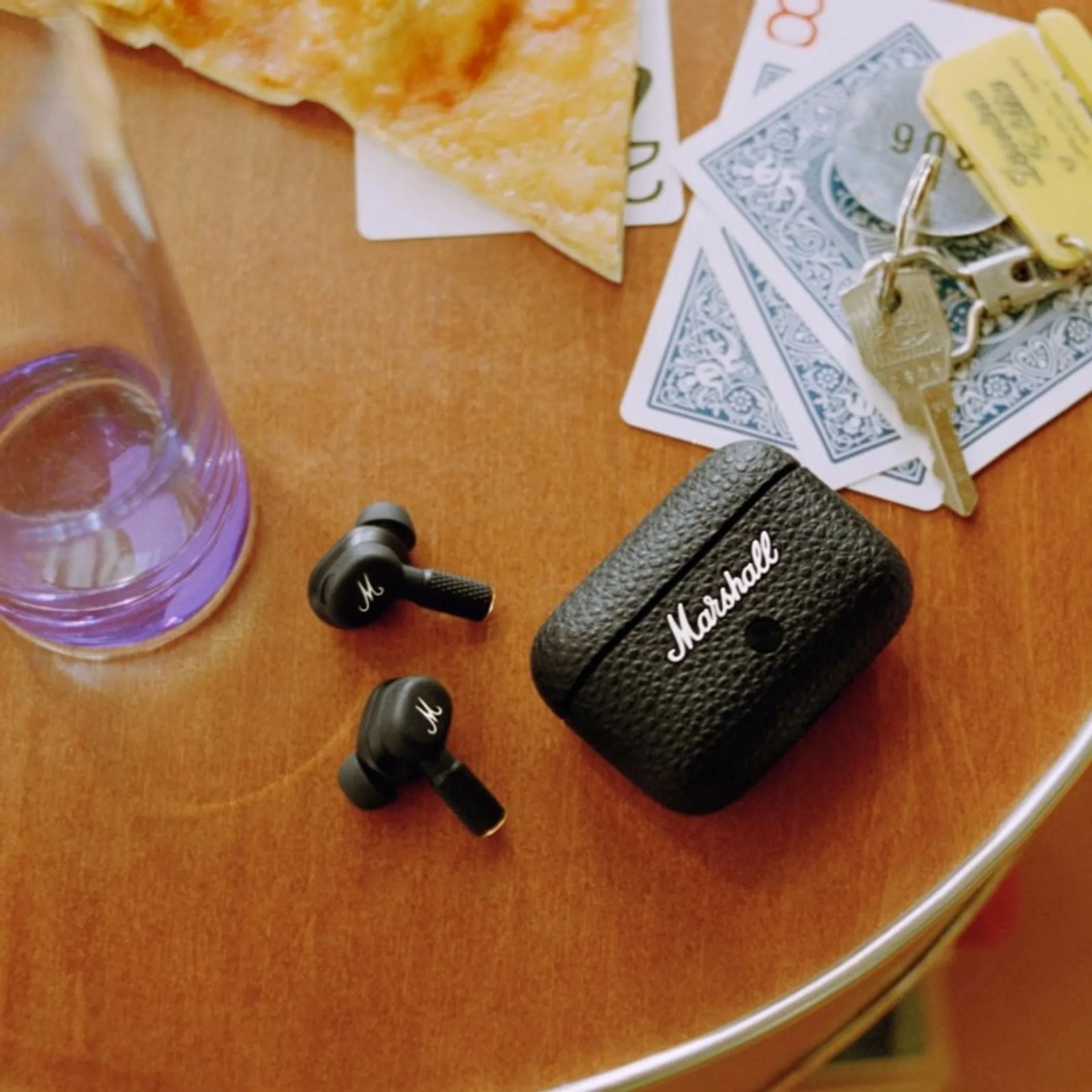 Marshall Motif II A.N.C. earbuds and leather-textured charging case on a wood table with pizza, playing cards, keys, and a p…