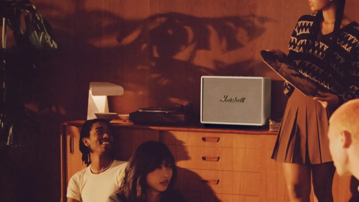 Friends in a warm wood-paneled room with cream Marshall speaker, turntable, and vinyl on a mid-century sideboard.