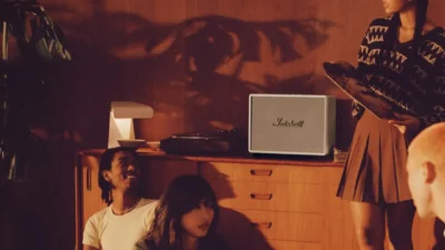 Friends in a warm wood-paneled room with cream Marshall speaker, turntable, and vinyl on a mid-century sideboard.