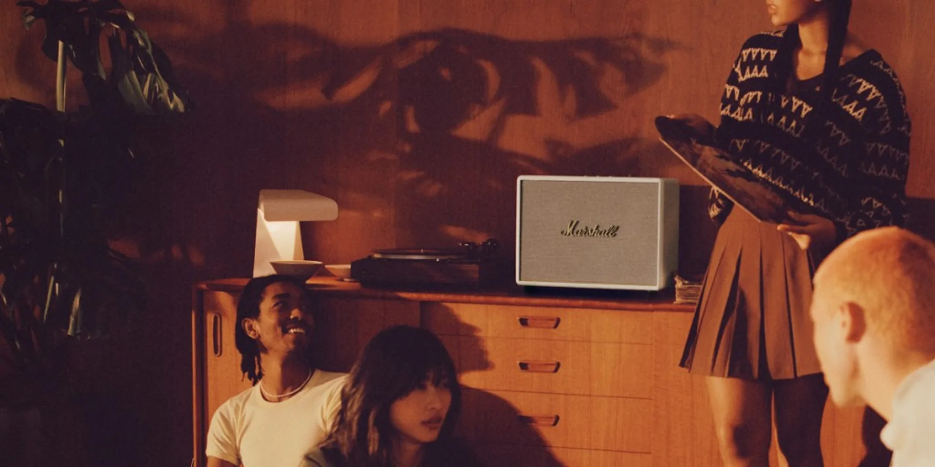 Friends in a warm wood-paneled room with cream Marshall speaker, turntable, and vinyl on a mid-century sideboard.