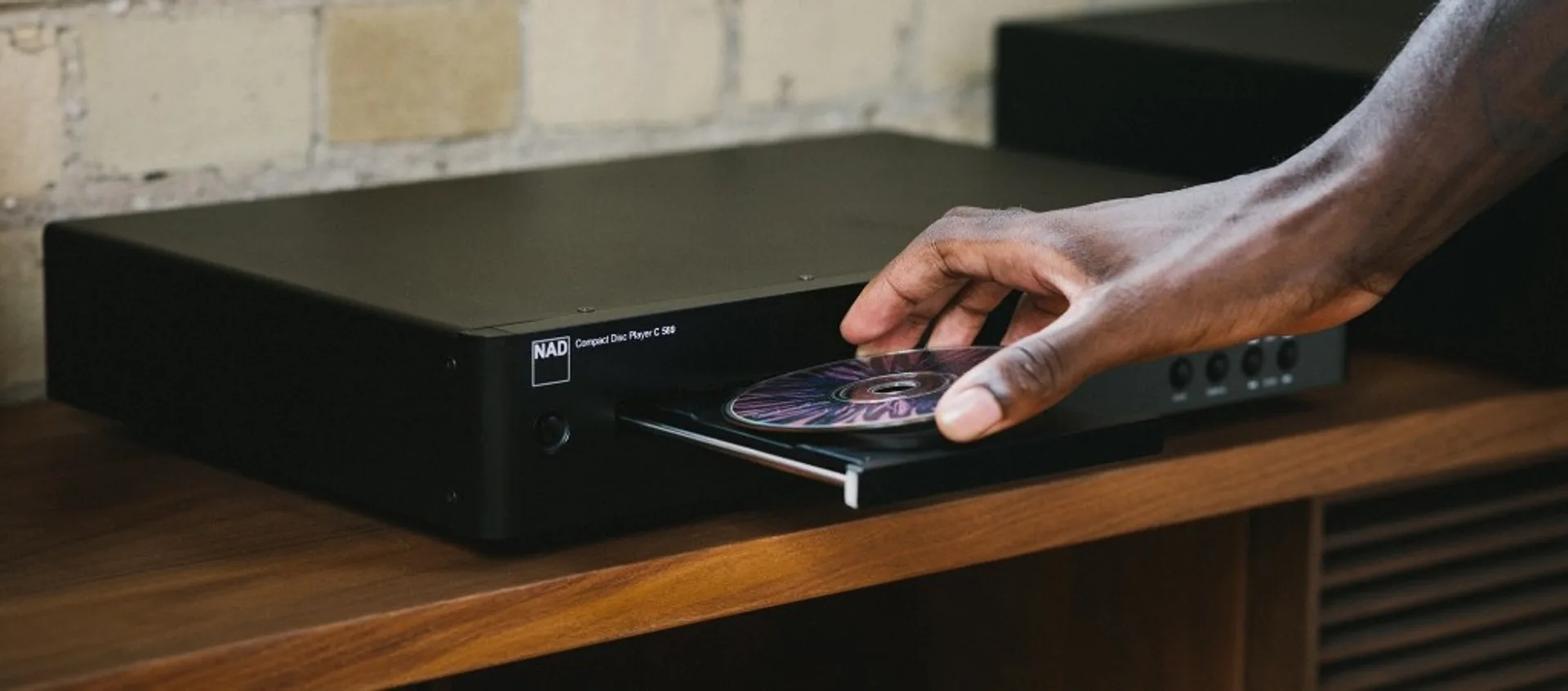 Lifestyle: arm placing colorful CD into NAD C 589 on polished wood credenza, soft brick wall background.