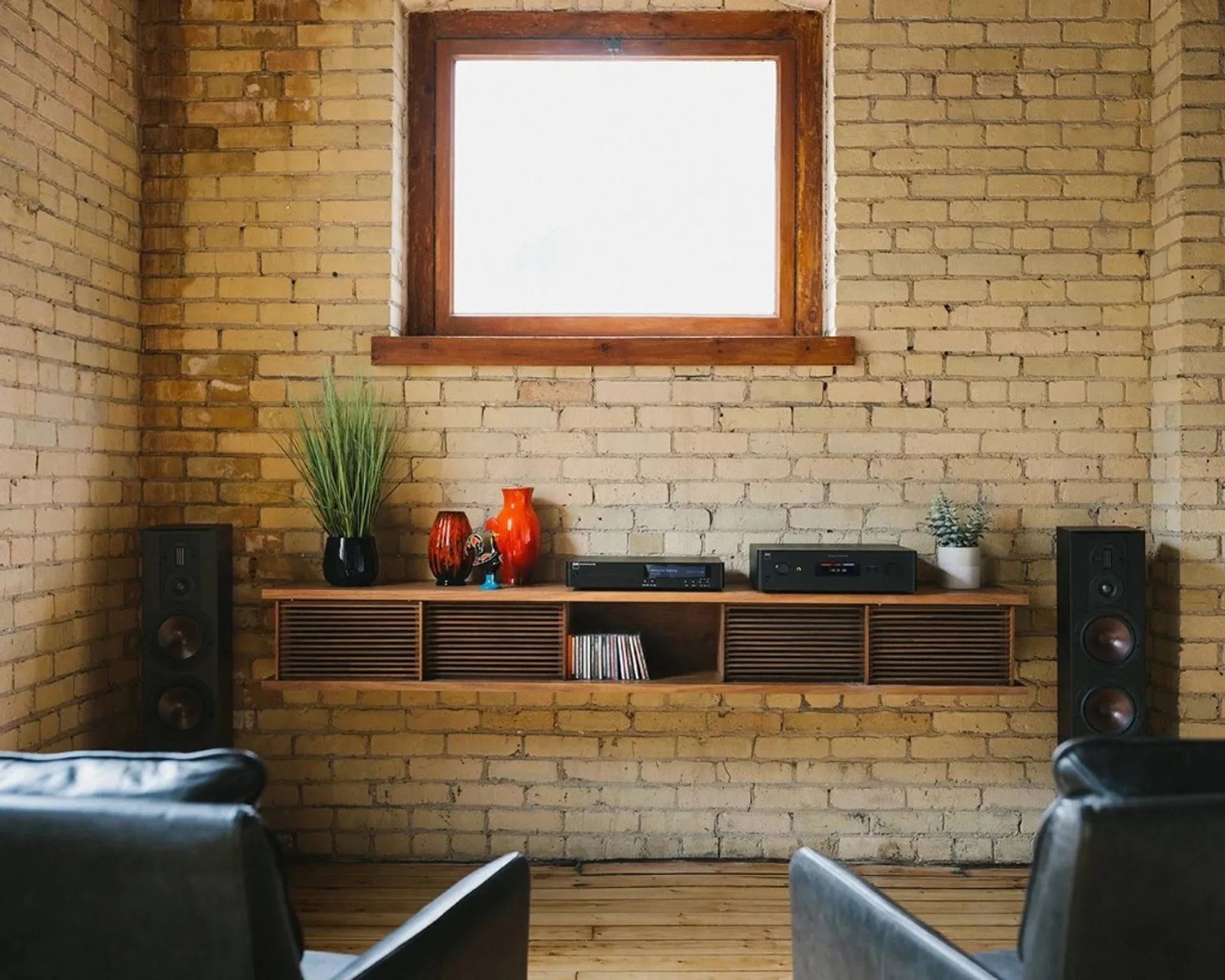 NAD C 589 CD player and integrated amplifier on floating wood console, floorstanding speakers, exposed brick wall and window, leather chairs foreground.