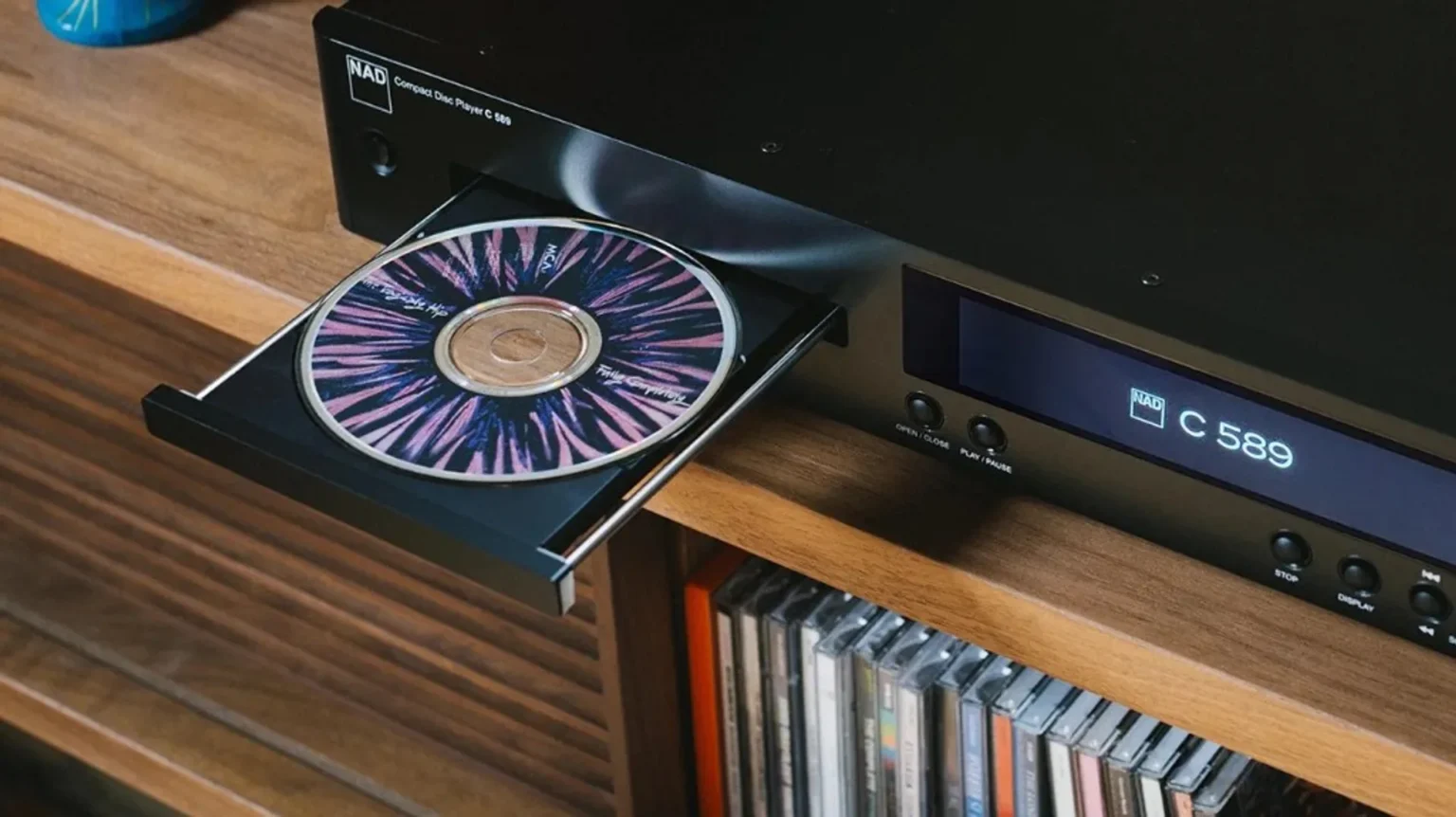 NAD C 589 compact disc player on light wood with open tray and purple burst CD, brick wall and orange vase, shallow depth of…