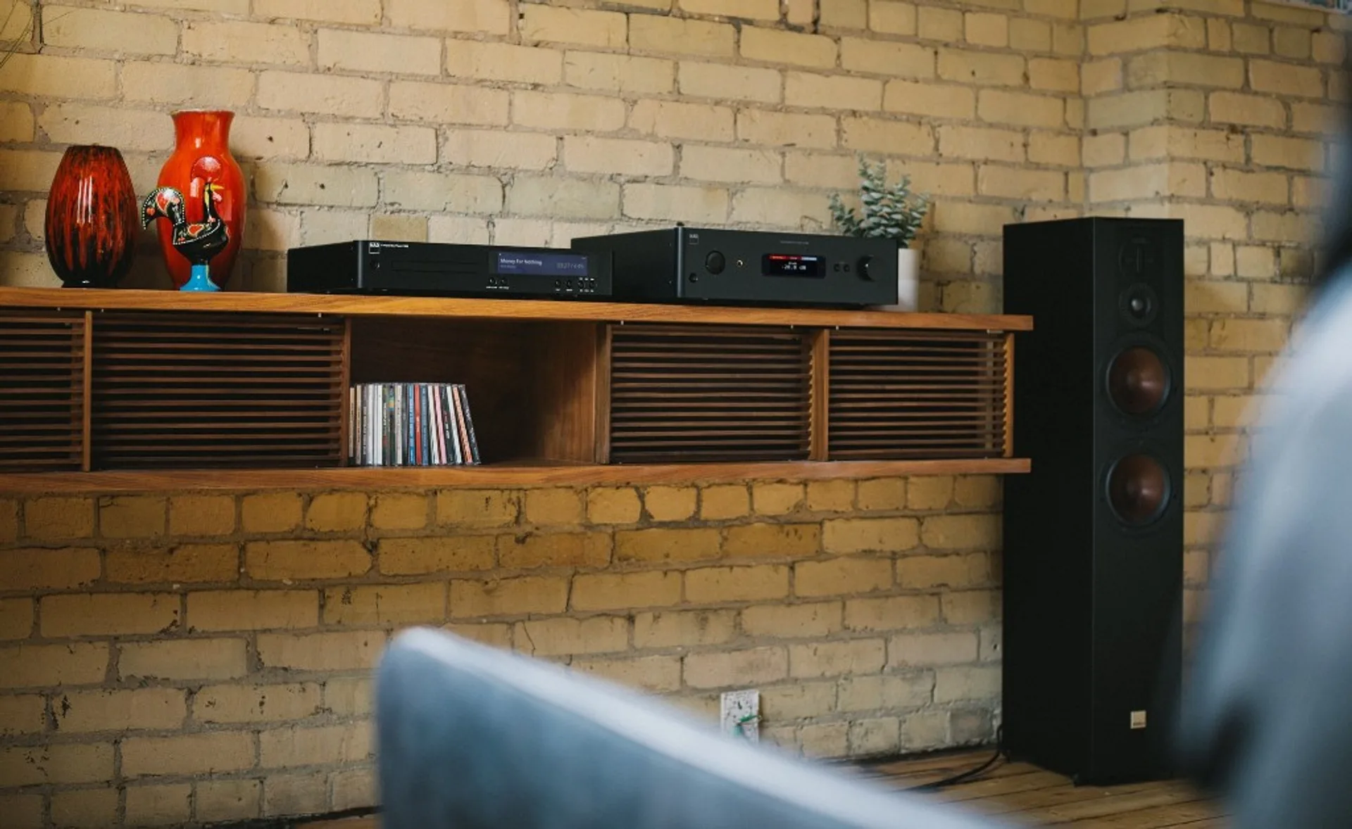 NAD C 589 and matching NAD integrated amplifier on wall-mounted wood shelf, brick wall, floorstanding speaker, decor vases.