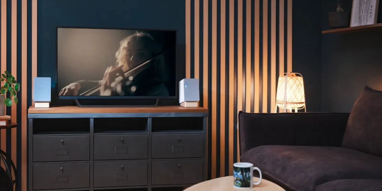 Onkyo Creator compact grey fabric speakers with wood bases on a dark media console flanking a TV in a teal and wood-slat liv…