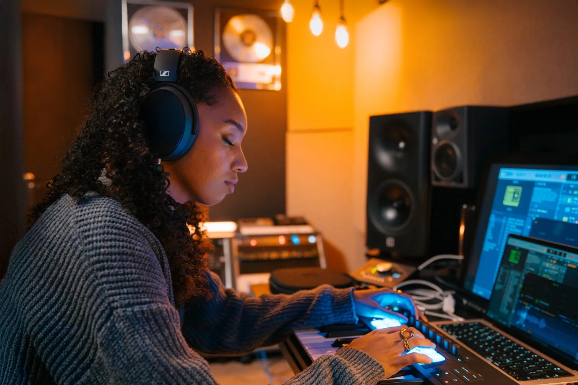 Woman producer in dim studio wearing Sennheiser headphones, hands on MIDI keyboard, DAW on laptop and monitor, warm filament lights.
