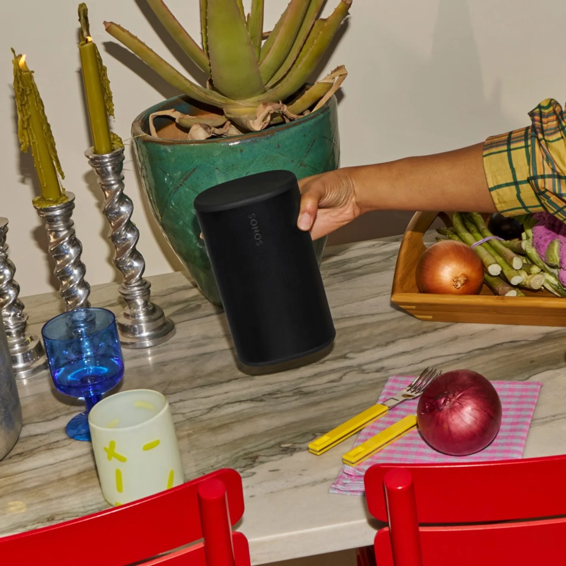 Hand carrying Sonos Play in a dining setting with plants, candles, and tableware, illustrating portable use.