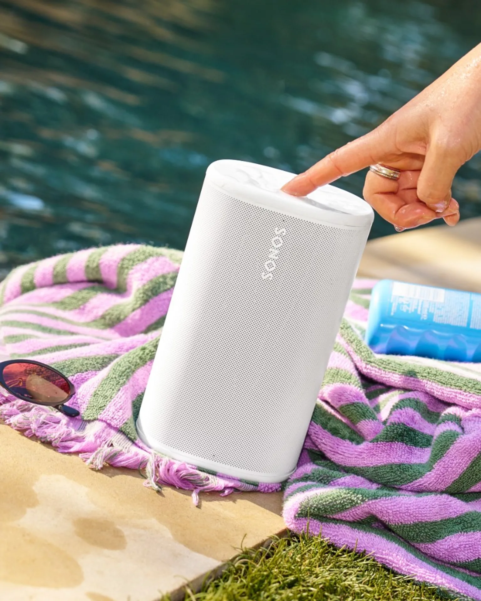 White Sonos Play at poolside on a towel with a hand touching top controls, illustrating splash-ready portable use.