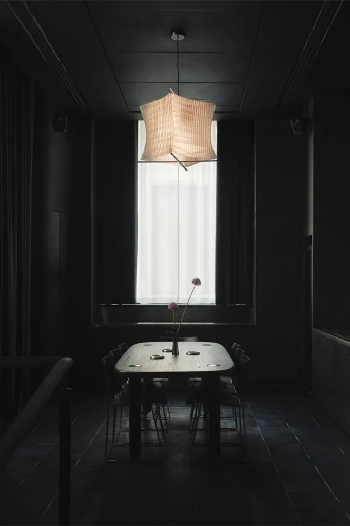Moody CAKE Architecture dining space with oval wood table, pleated pendant lamp, and bright curtained window.