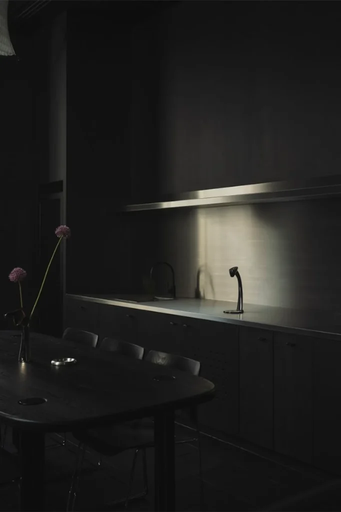 Minimal dark kitchen and dining area with stainless backsplash, black cabinetry, and purple flowers on wood table.