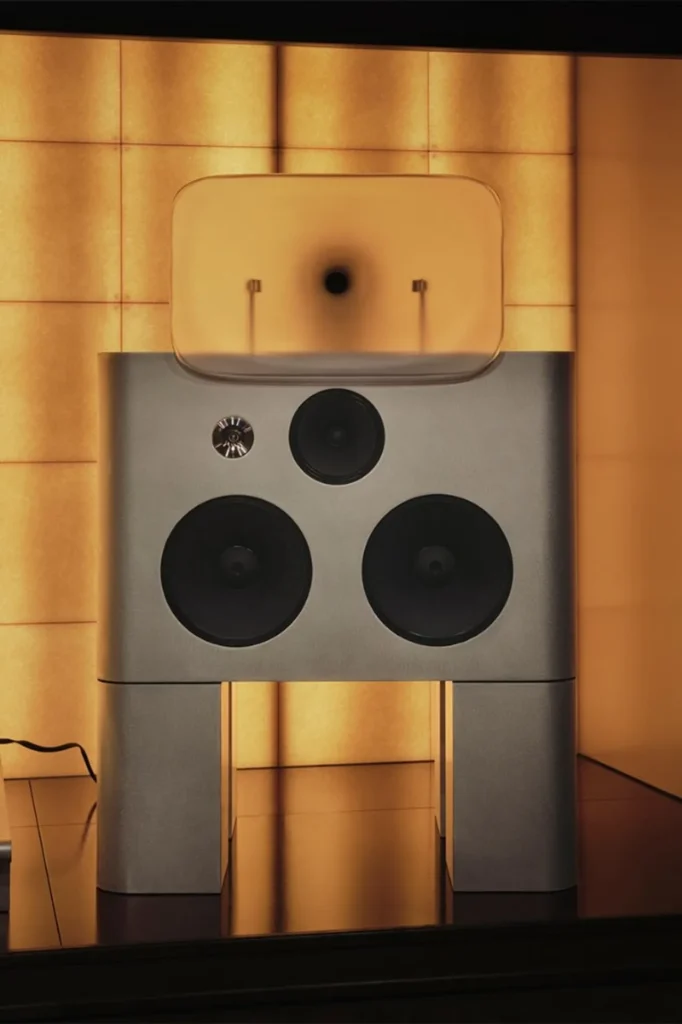 Full view of silver Friendly Pressure floor speaker with twin pedestal legs, drivers, and resin horn before glowing grid wal…