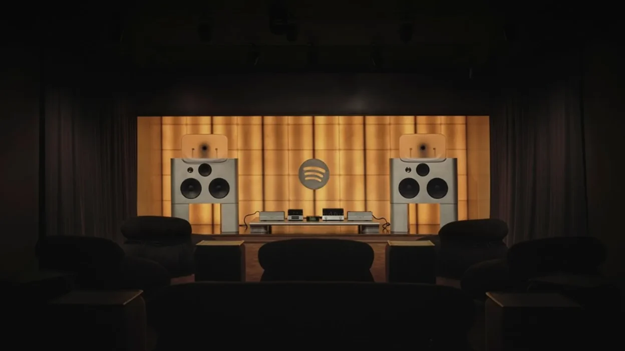 Wide shot of Spotify Listening Lounge stage with twin large speakers, Spotify logo on golden backlit wall, and audience seat…