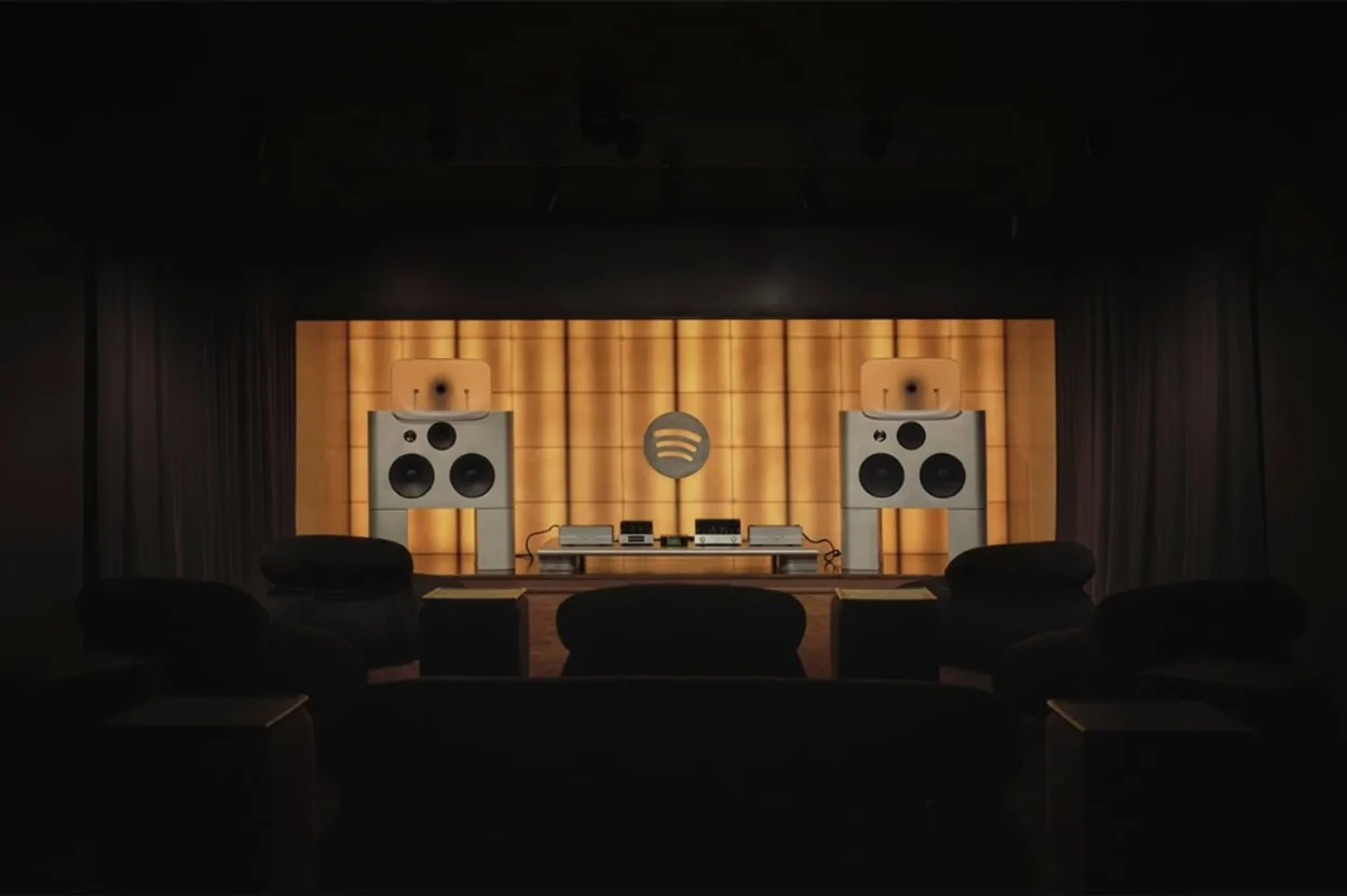Wide shot of Spotify Listening Lounge stage with twin large speakers, Spotify logo on golden backlit wall, and audience seat…