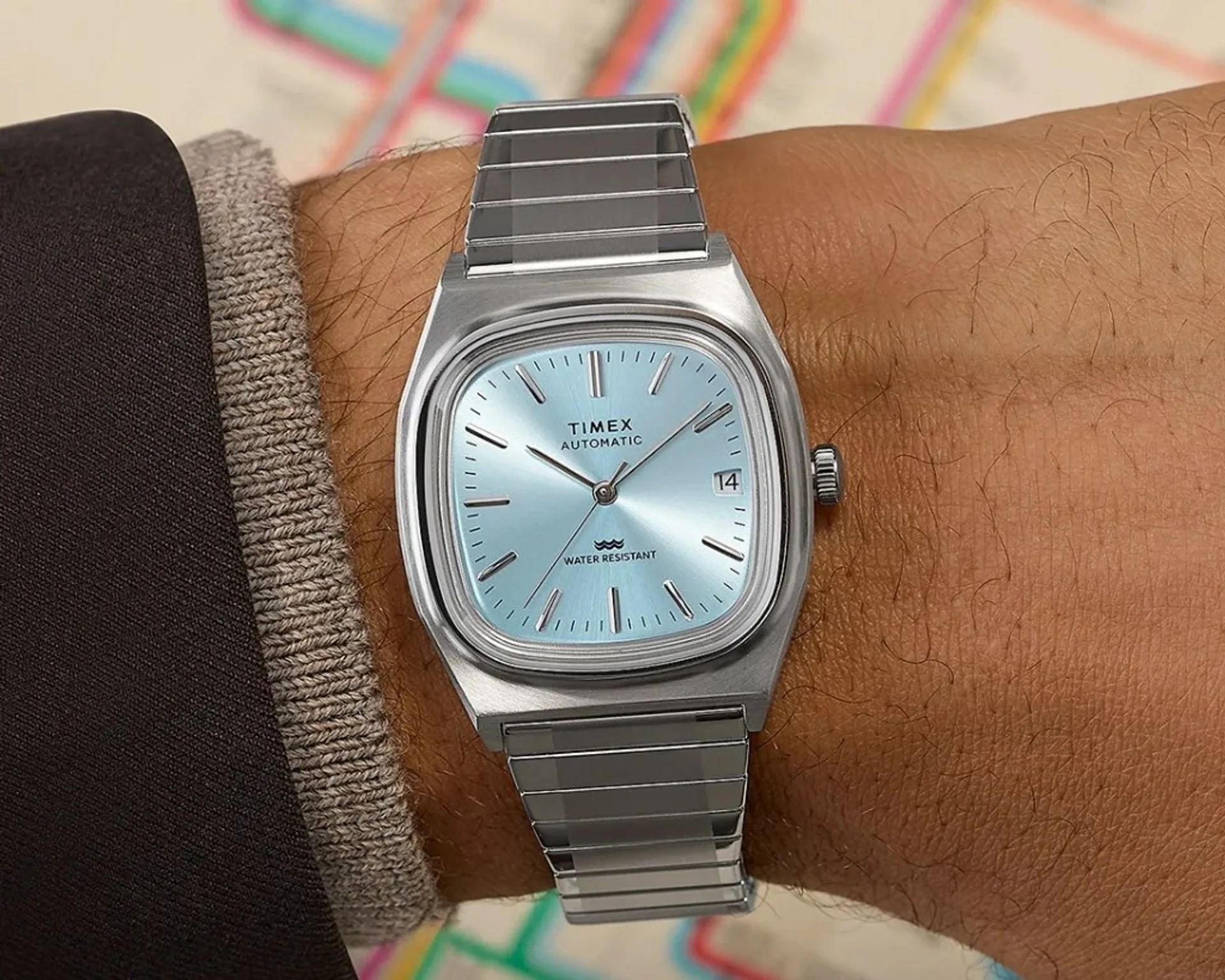 Timex Automatic 1983 E Line on wrist: ice blue sunburst dial, stainless cushion case, integrated steel bracelet, blurred map background.
