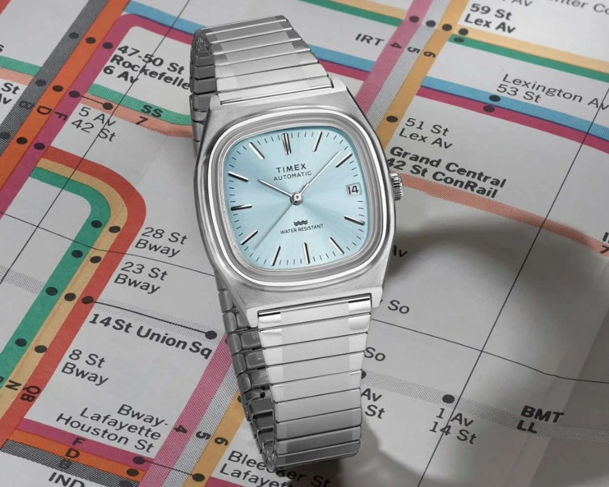Timex Automatic 1983 E Line with ice blue dial and date at three o'clock, stainless expansion bracelet on a colorful metro map.