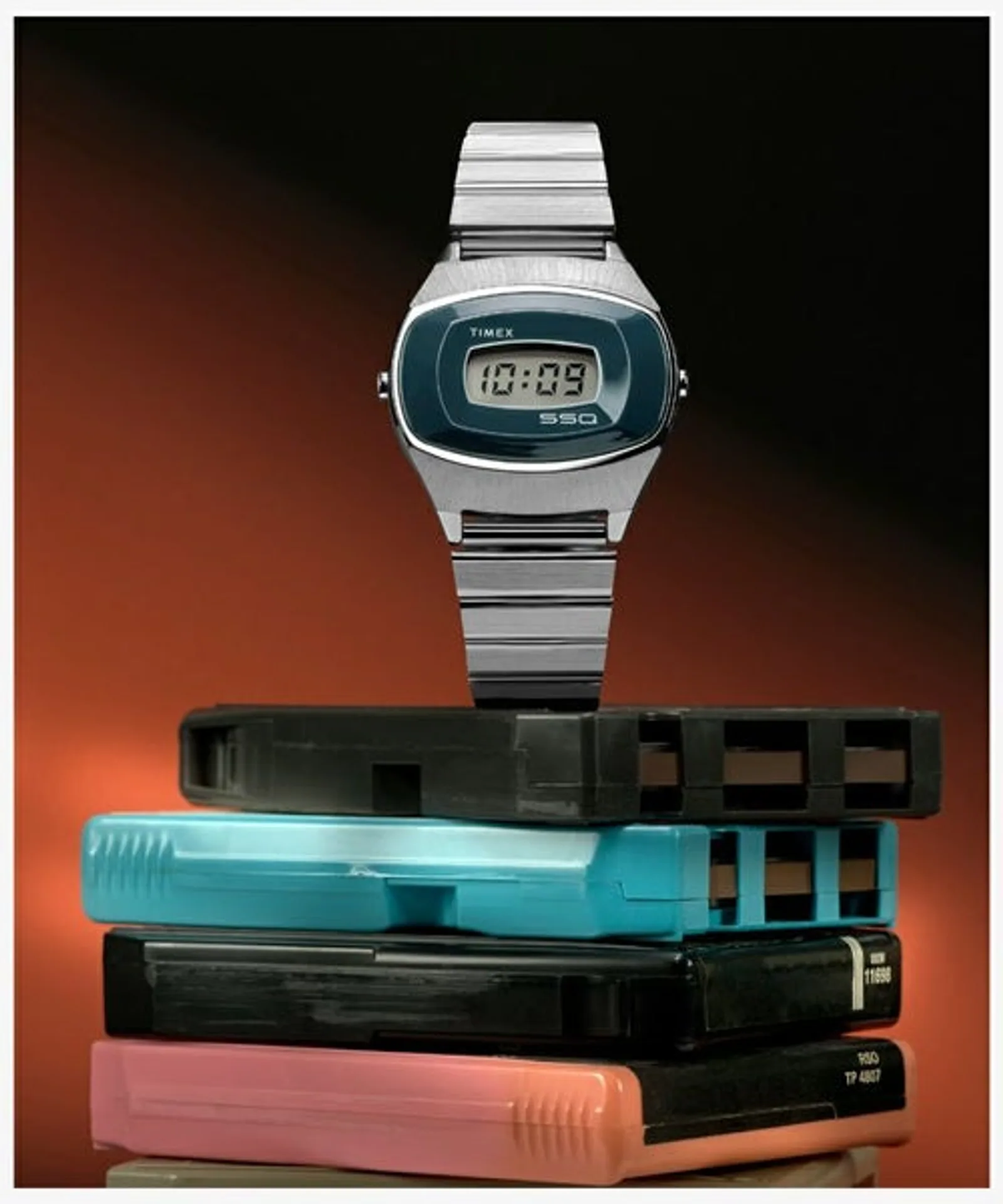 Timex SSQ digital watch on a stack of colorful 8-track cartridges against a warm orange-to-black gradient, retro 1970s styli…