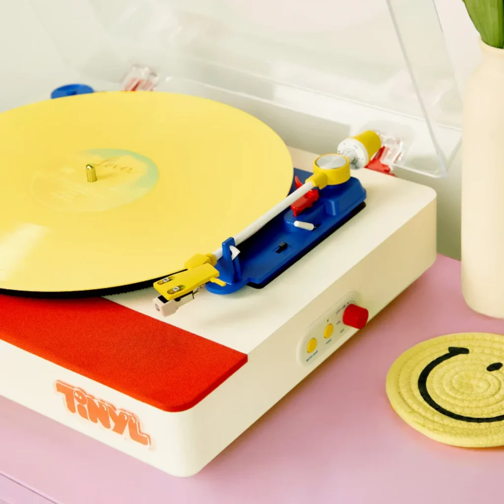 Close-up of Tinyl Play Pro on a pastel pink surface with yellow vinyl, red volume knob, and yellow PHONO and BT buttons besi…