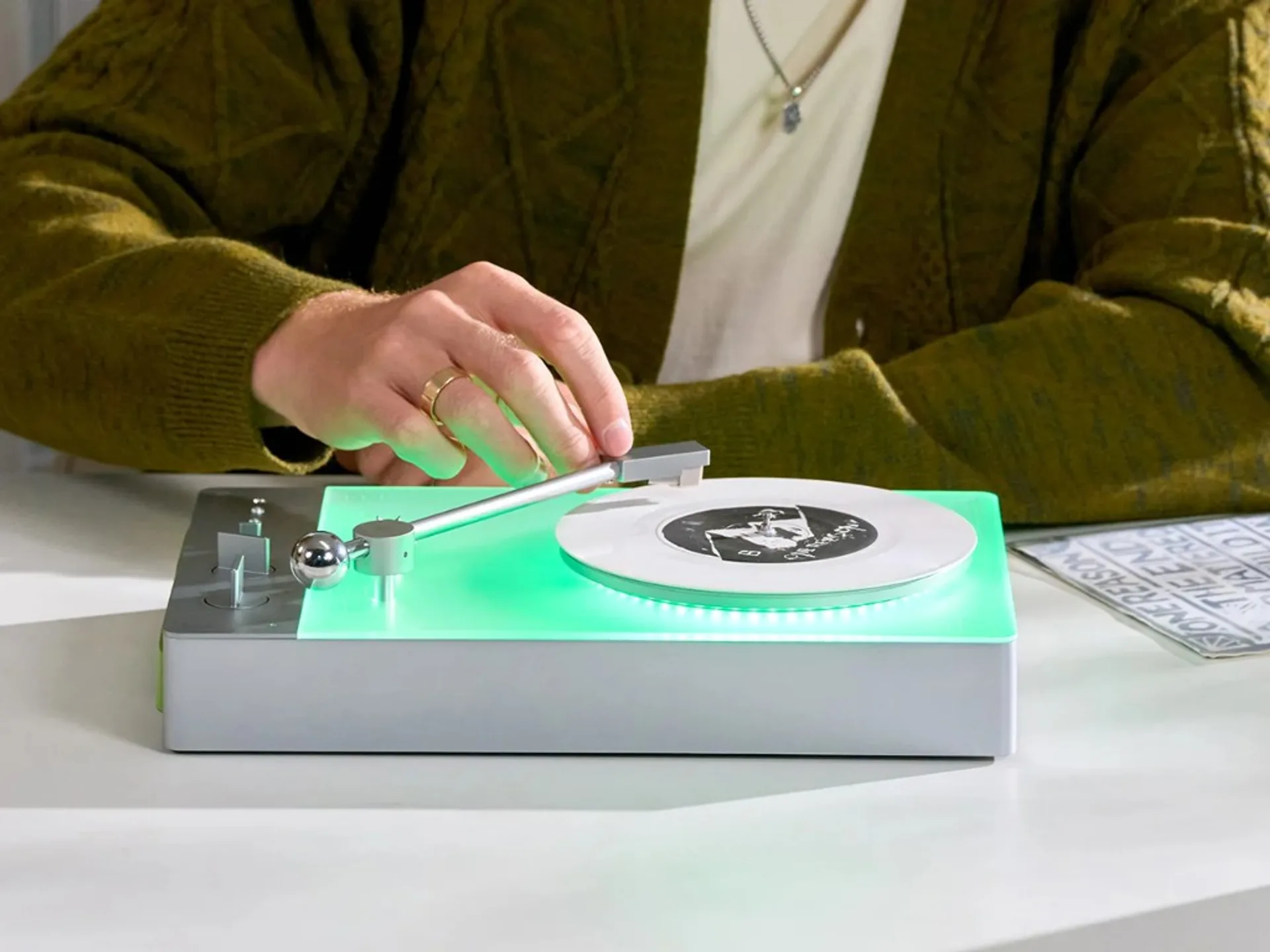 Person lowering the tonearm on a Trettitre TTT-LP3 wireless turntable with mint-green backlit translucent top plate, silver…