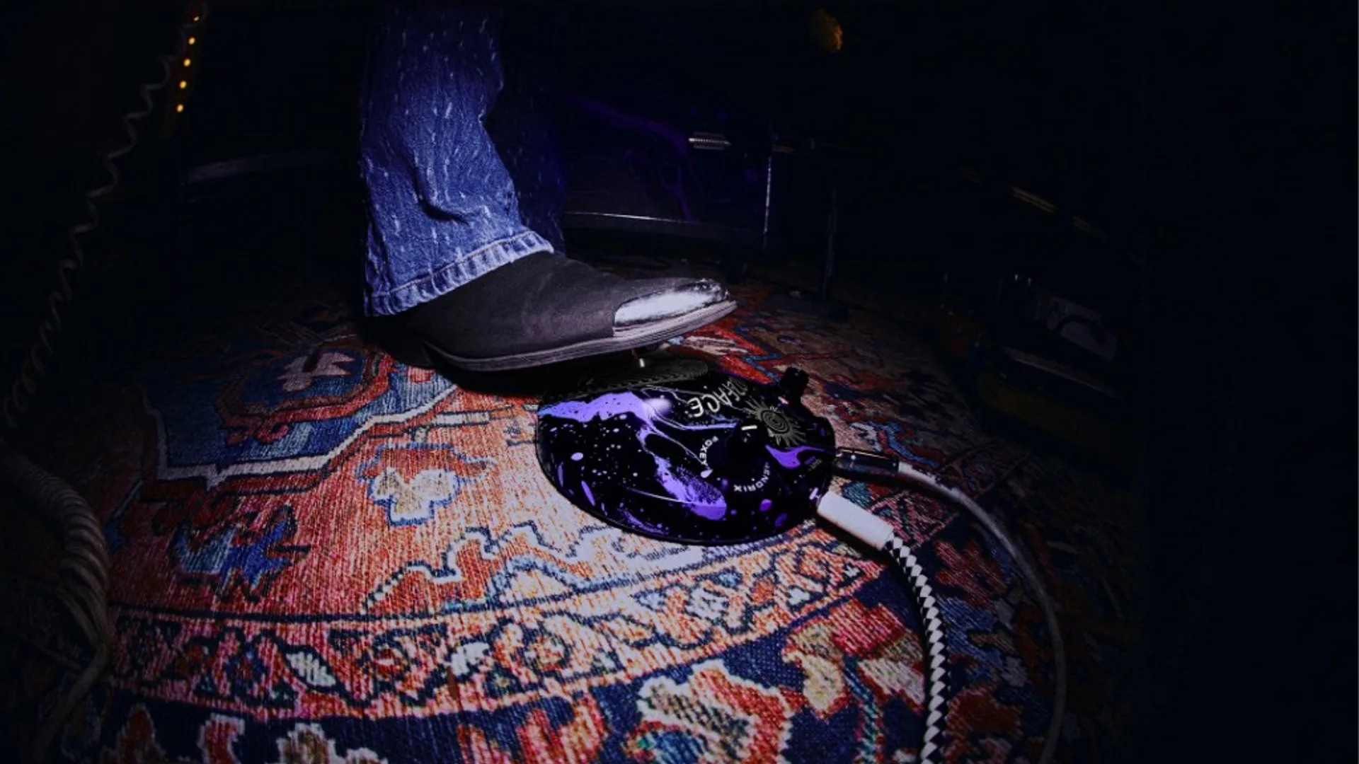 Wide-angle shot of a boot pressing a purple and black Hendrix Fuzz Face guitar pedal on a patterned rug with braided instrument cable.