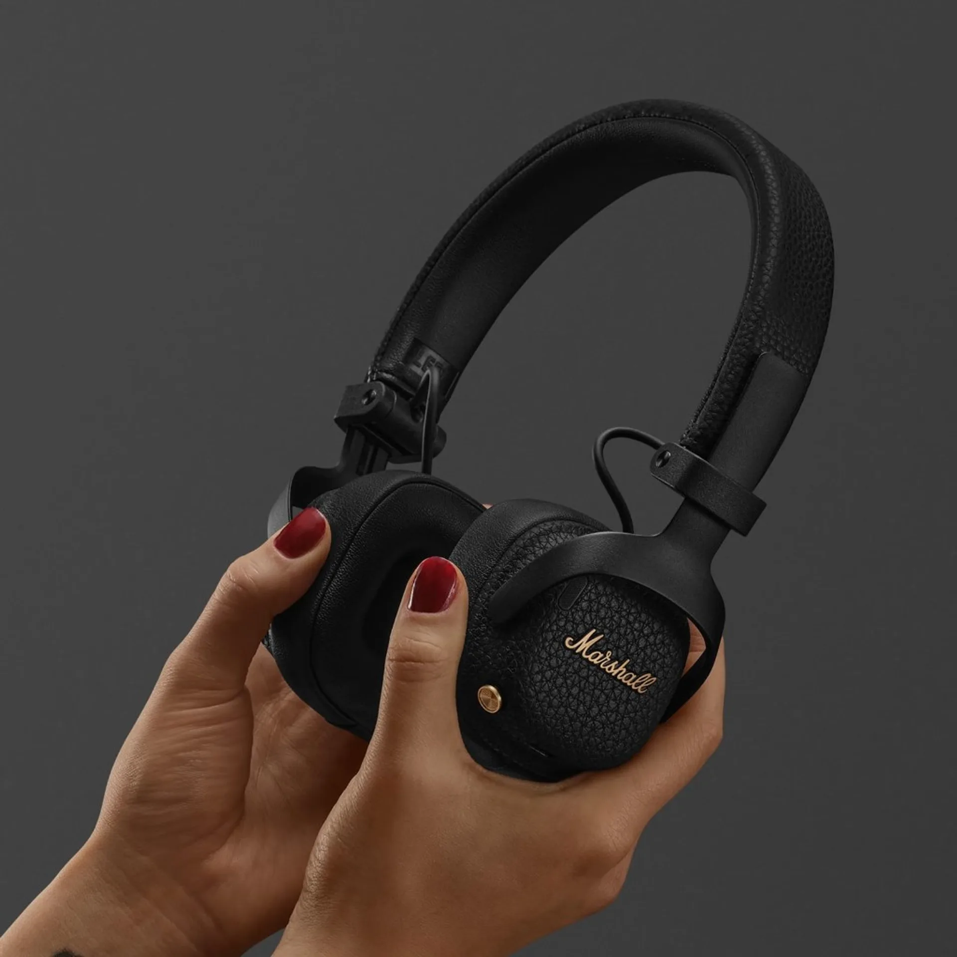 Hand with red nail polish holding compact Marshall Milton ANC on-ear headphones showing gold logo and textured ear cup.