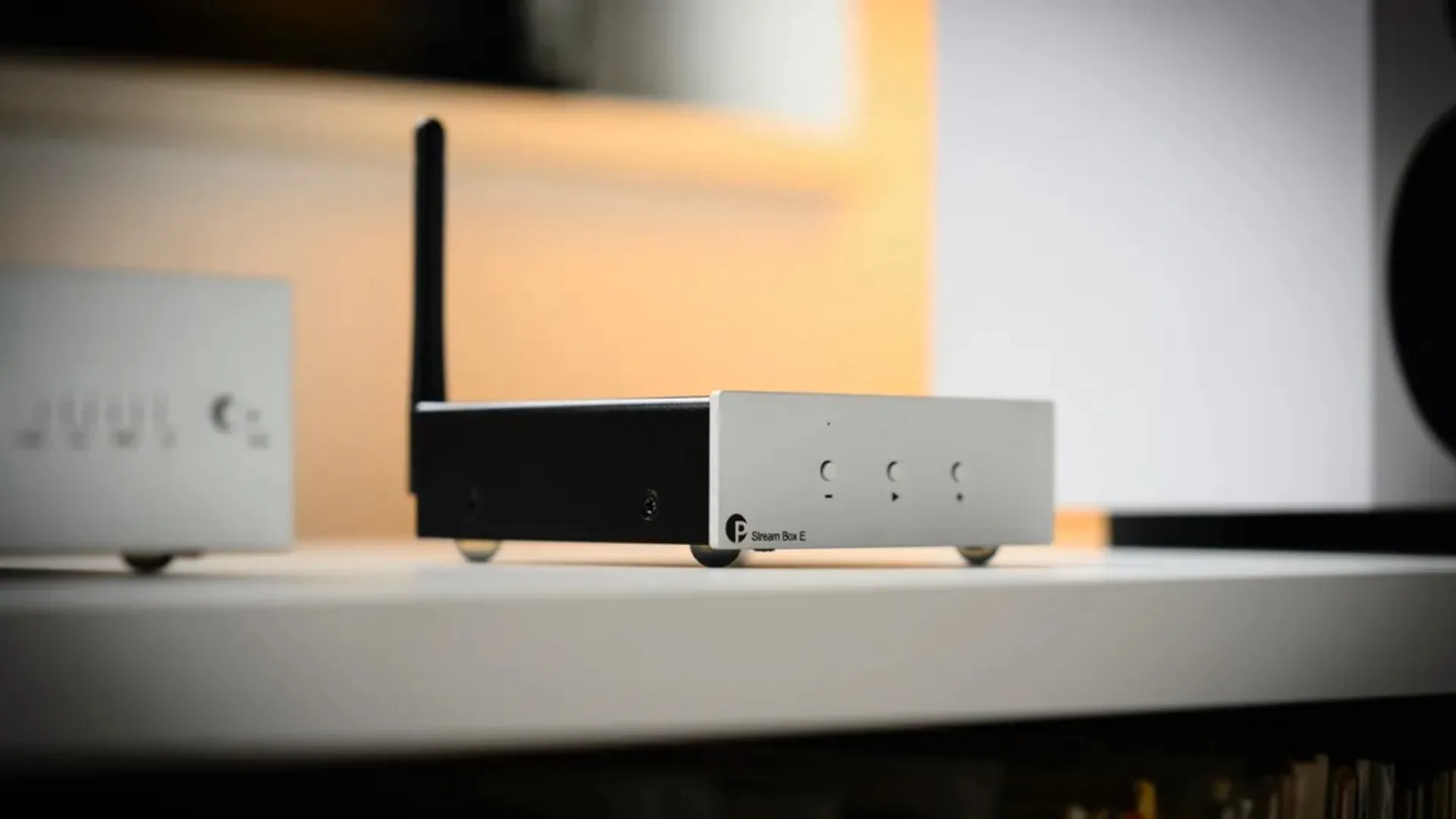 Pro-Ject Stream Box E and matching compact audio box on a white desk next to a smartphone displaying Spotify music playback.
