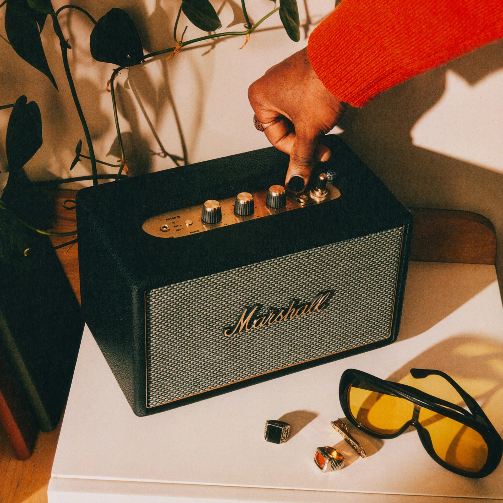 Hand flipping the brass power toggle on a Marshall Acton IV Black speaker with sunglasses and rings on a white surface.